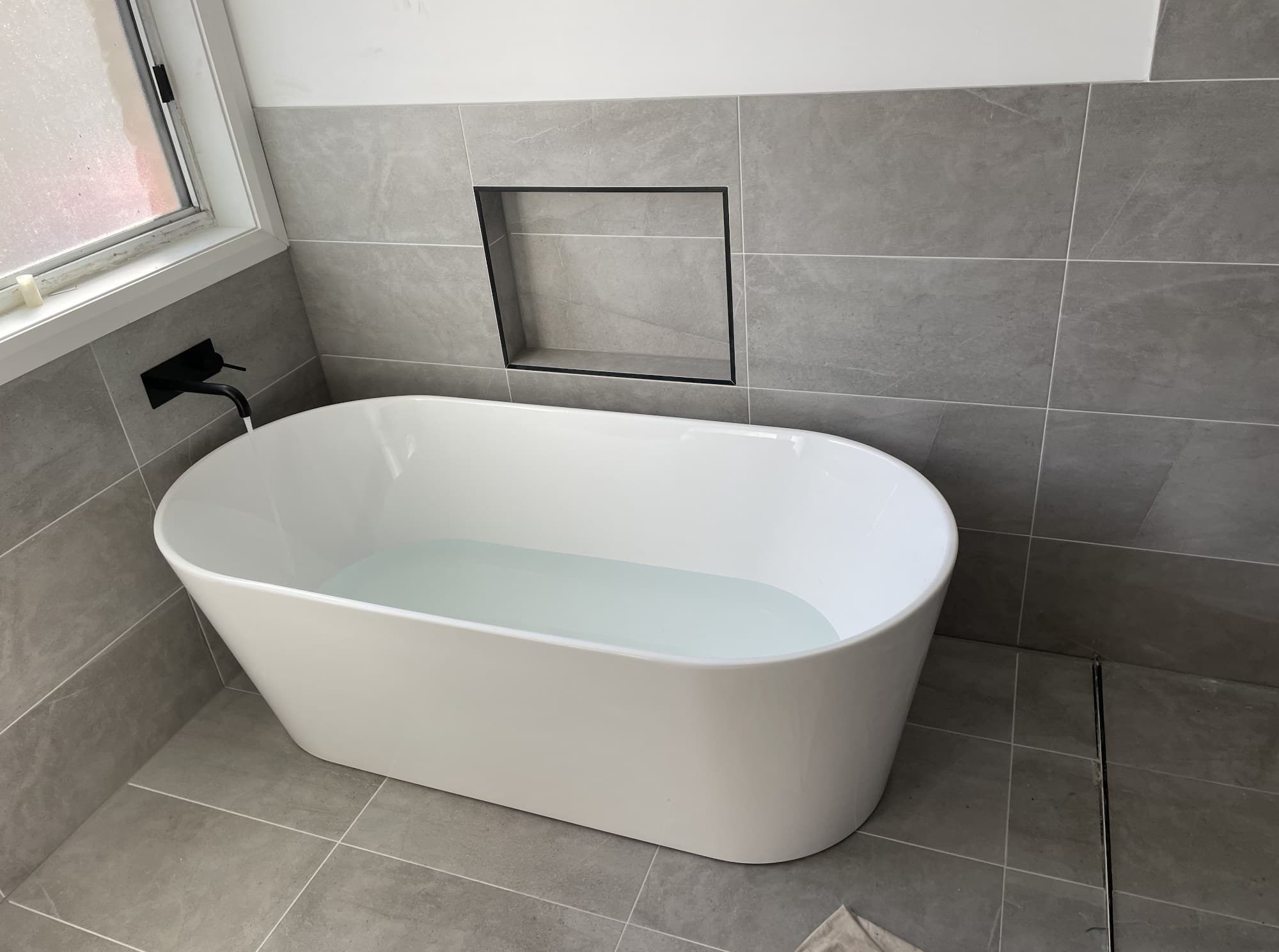 Freestanding bath plumbed in during a bathroom renovation, with matte black tapware and floor-to-ceiling tiling, Kiama NSW